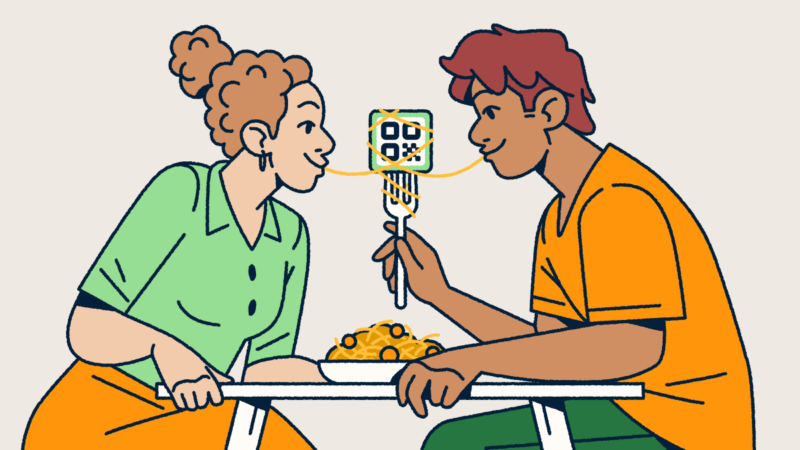 A couple sitting at a table sharing a bowl of spaghetti and meatballs. One partner is holding a fork with a QR Code on it and a piece of spaghetti wrapped around it. They each have one end of the noodle in their mouth.