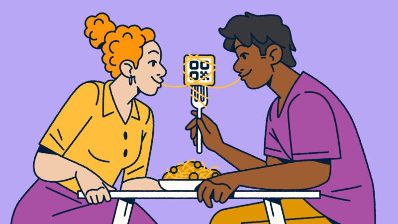 A couple sitting at a table sharing a bowl of spaghetti and meatballs. One partner is holding a fork with a QR Code on it and a piece of spaghetti wrapped around it. They each have one end of the noodle in their mouth.